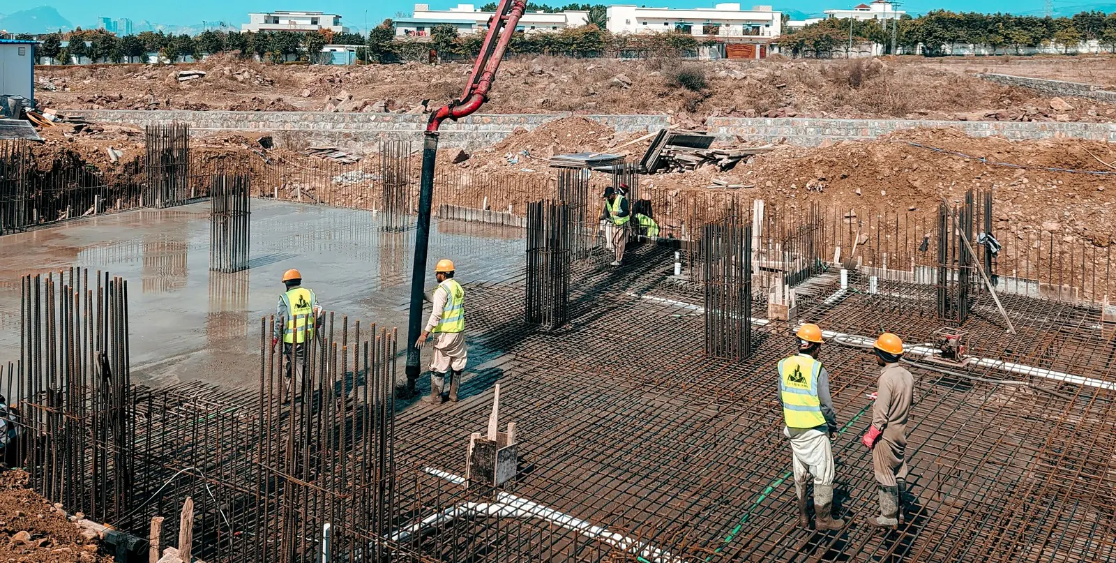 A slab on grade part way through a pump pour: the rebar mat is set and tied with column starters standing, wet concrete covers the left half, and four workers in hi-vis stand on the mat.