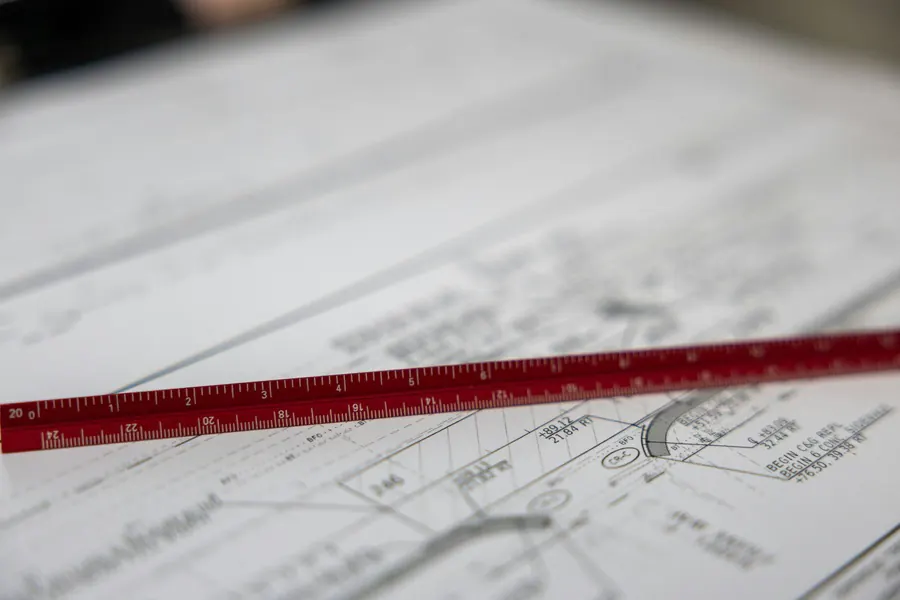 An architect's triangular scale rule lying across a civil plan sheet, with curb-and-gutter and sidewalk callouts and station offsets visible under it.